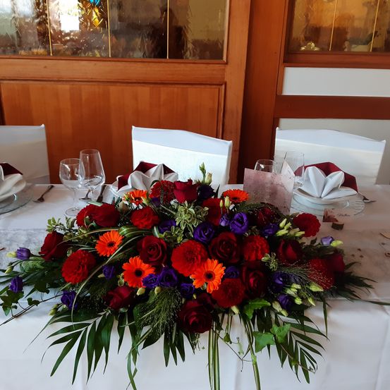 Eine Blumenarrangement mit roten, lila und orangefarbenen Blumen steht auf einem Tisch. Vier Weingläser, ein weißer Teller und eine Serviette liegen auf dem Tisch. Die Stühle haben weiße Stoffbezüge. Der Tisch ist mit einem weißen Tuch bedeckt.