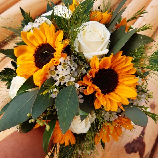 Ein Nahaufnahme einer Blumenarrangements zeigt zwei Sonnenblumen und eine weiße Rose, umgeben von grünen Blättern und weißen Blüten.