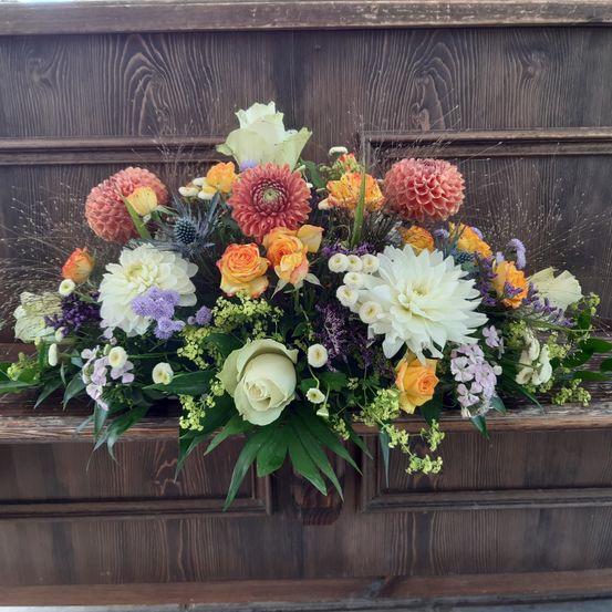 Ein farbenfrohes Blumenarrangement mit weißen, orangen und lilafarbenen Blumen steht auf einer Holzoberfläche. Das Bouquet enthält Rosen, Dahlien und Grünpflanzen.