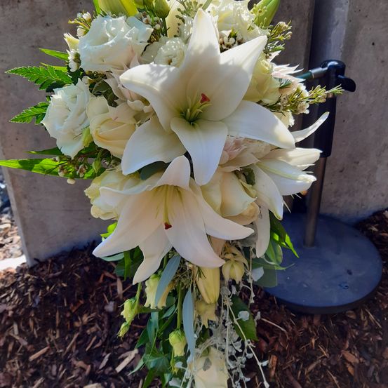 Ein Strauß weißer Blumen, einschließlich Lilien und Rosen, mit grünen Blättern arrangiert, steht auf einem Ständer im Freien.
