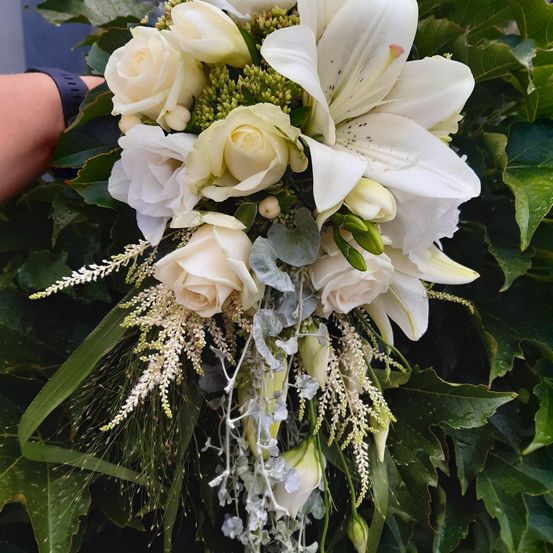 Ein Nahaufnahme eines Brautstraußes mit weißen Rosen, Lilien und Grünpflanzen, der von einer Hand gehalten wird.