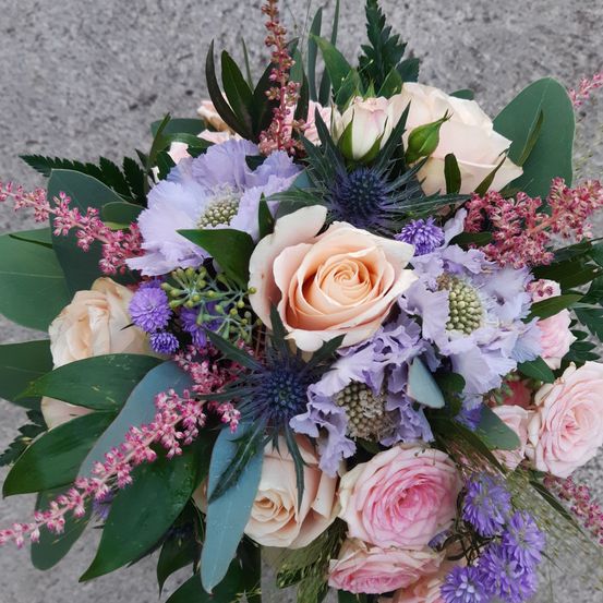 Ein Blumenstrauß mit verschiedenen Farben und Arten von Blumen, einschließlich Rosen, Lavendel und lila Blüten, vor einem grauen Hintergrund.
