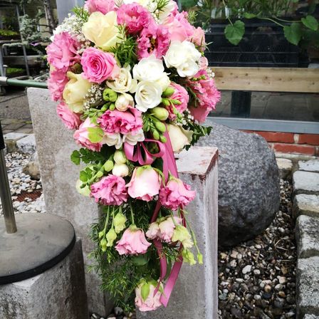 Ein Blumenarrangement mit rosa, weißen und cremefarbenen Rosen, geschmückt mit Grünpflanzen und mit einem rosa Band gebunden, ruht auf Betonblöcken im Freien.