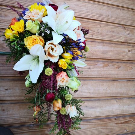 Ein farbenfroher Blumenstrauß mit Lilien, Rosen und verschiedenen farbenfrohen Blüten, arrangiert an einer Holzwand.
