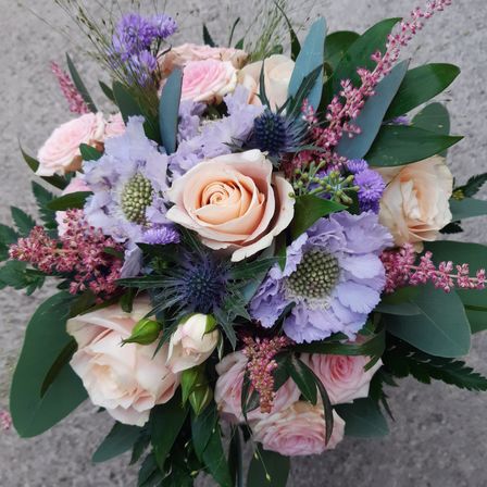 Ein farbenfroher Blumenstrauß mit einer Mischung aus rosa, weißen und lila Rosen, Lavendelblüten und Disteln, umgeben von grünen Blättern, ist in einer kreisförmigen Form auf einer grauen Oberfläche angeordnet.