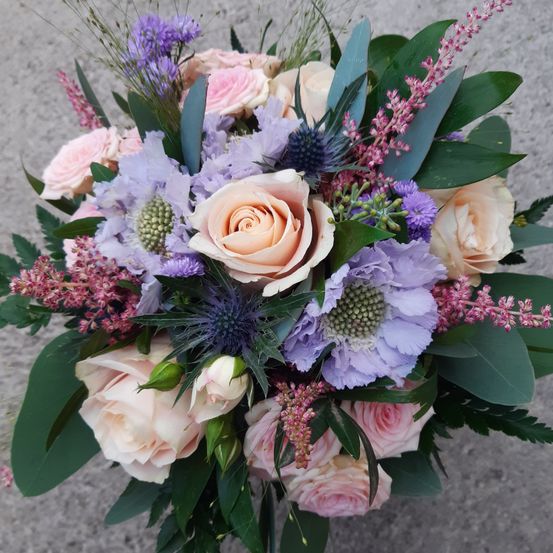 Ein farbenfroher Blumenstrauß mit einer Mischung aus rosa, weißen und lila Rosen, Lavendelblüten und Disteln, umgeben von grünen Blättern, ist in einer kreisförmigen Form auf einer grauen Oberfläche angeordnet.