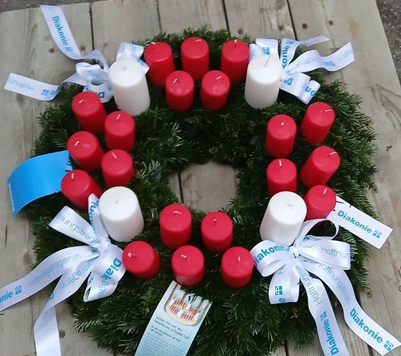 Ein Kranz mit roten und weißen Kerzen liegt auf einer Holzoberfläche. Er ist mit weißen Bändern und Fahnen geschmückt. Jede Kerze hat einen weißen Docht. Der Kranz liegt auf einer Holzoberfläche.