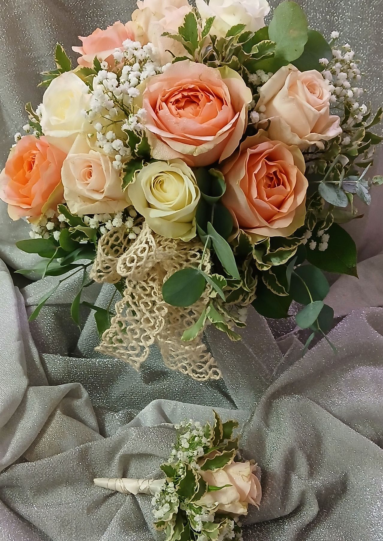 Ein Brautstrauß aus rosa und weißen Rosen mit Babys Atem und grünen Blättern, in Spitze gewickelt, auf einem silbernen Stoff.