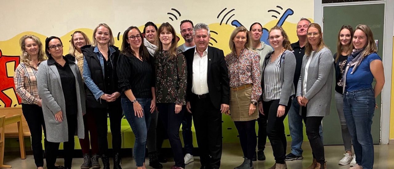 Eine Gruppe von Erwachsenen, sowohl Männer als auch Frauen, stehen in einem Raum mit einem farbenfrohen Wandbild, in lässiger Kleidung.