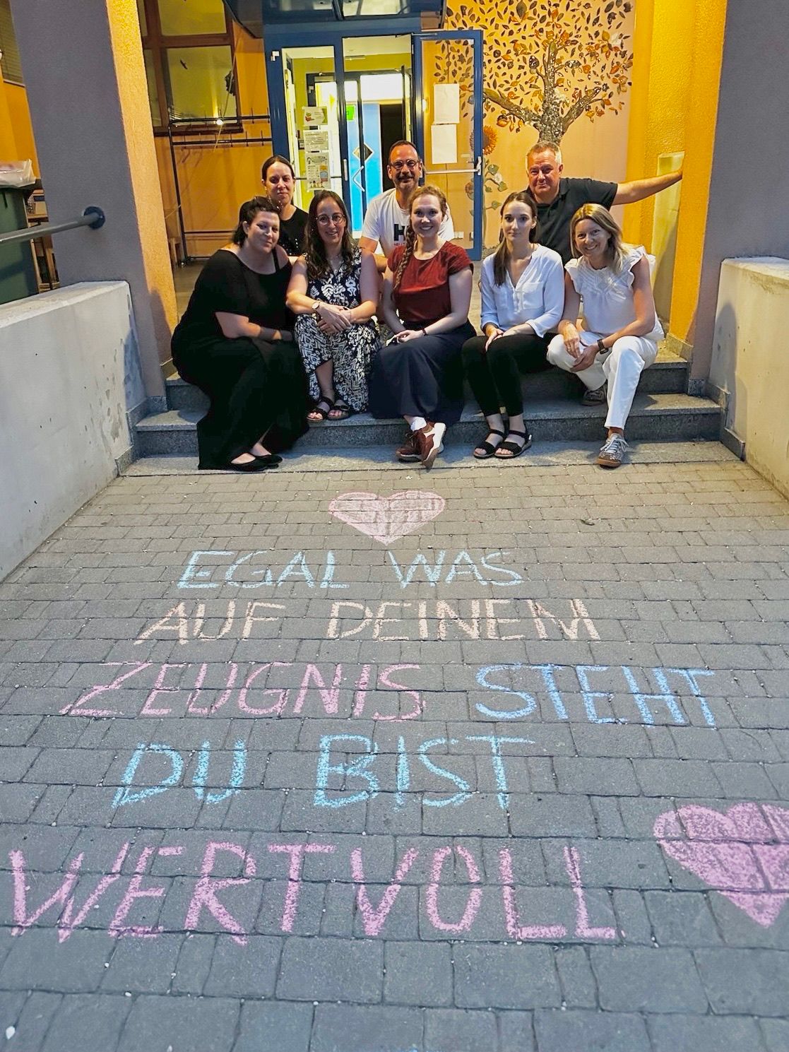 Eine Gruppe von Menschen sitzt auf Stufen mit einer Kreidebotschaft auf dem Boden, die 'Egal was auf deinem Zeugnis steht, du bist wertvoll' lautet.