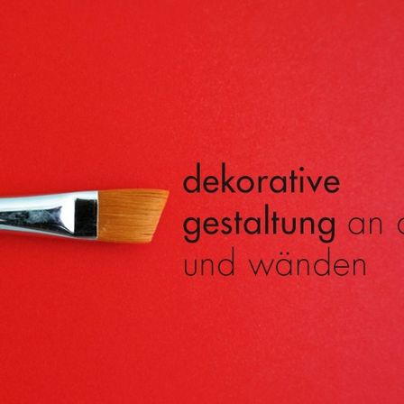 Ein Pinsel auf rotem Hintergrund mit Text, der 'dekorative Gestaltung an Decken und Wänden' lautet. Der Pinsel hat einen silbernen Griff und eine hölzerne Spitze.