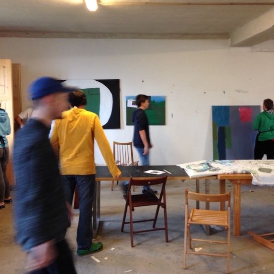 Ein Kunststudio mit mehreren Personen. Eine Person steht an einem langen Tisch, eine andere lehnt sich mit Gemälden an eine Wand. Stühle und Tische sind verstreut.