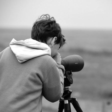 Ein Junge in einem Hoodie schaut durch ein auf einem Stativ montiertes Teleskop, möglicherweise beobachtet er den Ozean.