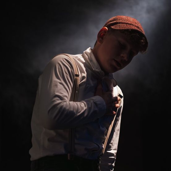 Ein junger Mann in einem weißen Hemd und mit Hosenträgern, der einen Hut trägt, möglicherweise schläft, auf einer Bühne bei schwacher Beleuchtung.