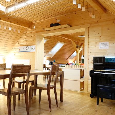 Ein hölzerner Raum mit Tisch und Stühlen. Ein Klavier steht an einer Wand. Ein Schreibtisch mit Buntstiften und anderen Gegenständen befindet sich neben dem Klavier. Der Raum hat Holzwände und Decke mit Beleuchtung.