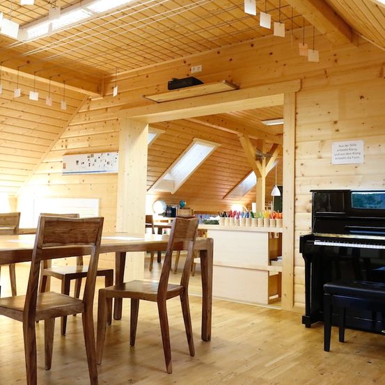 Ein hölzerner Raum mit Tisch und Stühlen. Ein Klavier steht an einer Wand. Ein Schreibtisch mit Buntstiften und anderen Gegenständen befindet sich neben dem Klavier. Der Raum hat Holzwände und Decke mit Beleuchtung.