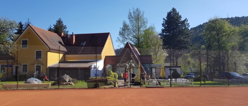 Ein Tennisplatz mit einem gelben Gebäude im Hintergrund. Mehrere Personen stehen in der Nähe des Eingangs, und ein Auto ist in der Ferne geparkt.