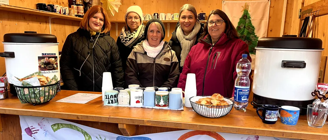 Fünf Frauen stehen hinter einer Theke in einem kleinen Holzraum. Sie lächeln und scheinen für ein Foto zu posieren. Die Theke hat verschiedene Gegenstände darauf, einschließlich Tassen, Flaschen und ein Banner mit dem Text '65 Stegersbach'.
