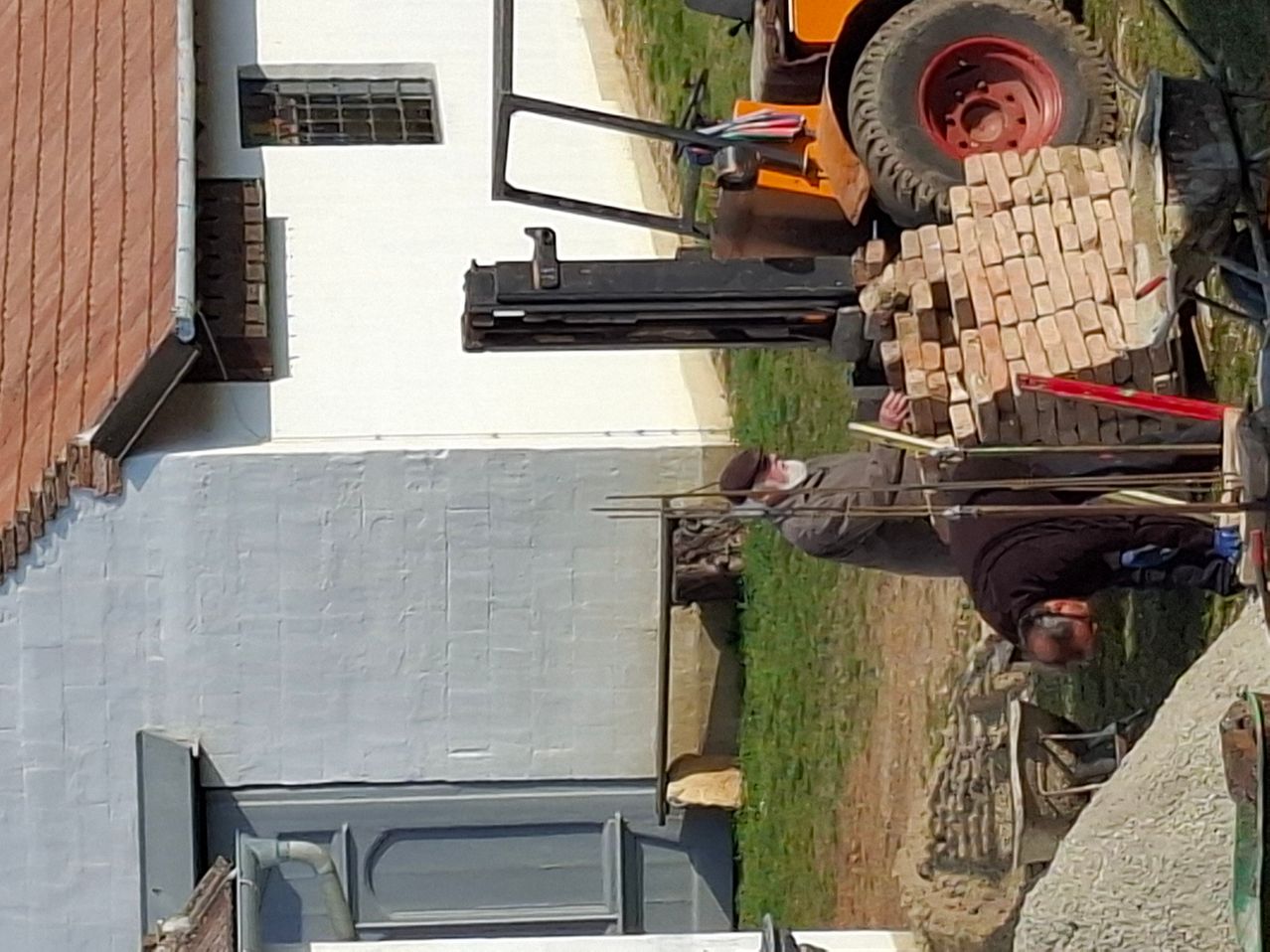 Eine Vogelperspektive eines Gebäudes zeigt zwei Männer in der Nähe eines Gabelstaplers mit einem Stapel Ziegel. Das Gebäude hat eine weiße Wand und einen Backsteinkamin.