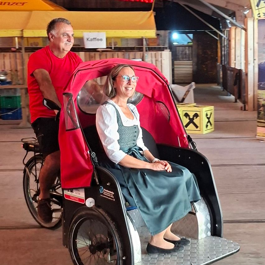 Ein Mann und eine Frau fahren einen roten Rikscha. Der Mann tritt in die Pedale, während die Frau vorne sitzt. Sie lächeln beide.