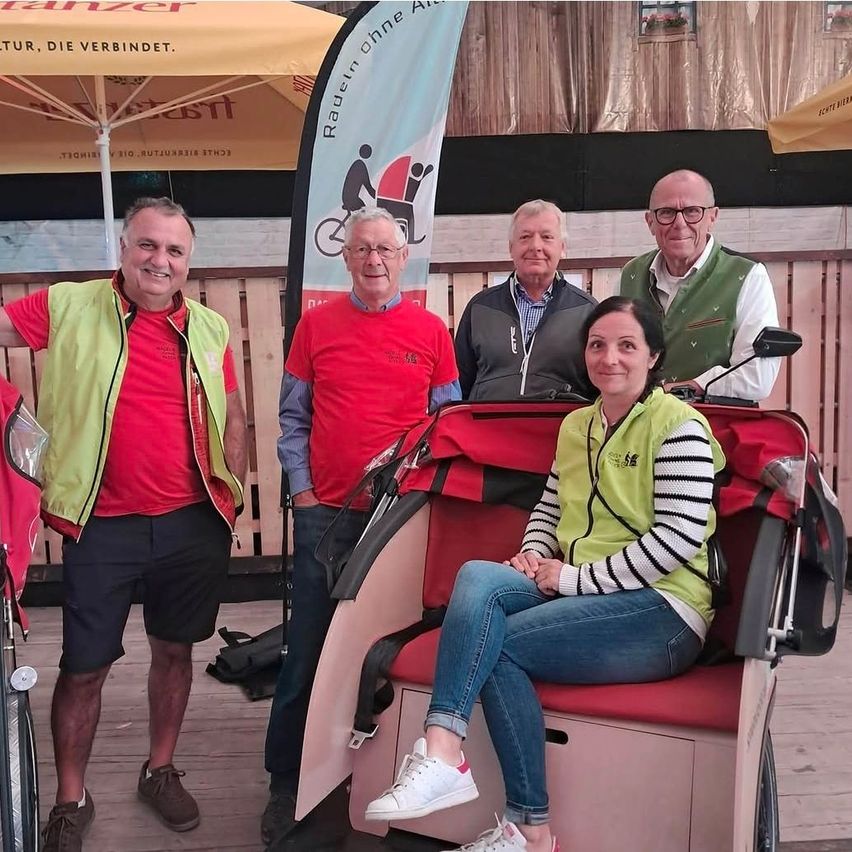 Fünf Personen stehen neben einem roten Dreirad und lächeln für ein Foto. Zwei Männer tragen rote Hemden, einer trägt eine grüne Weste und zwei tragen eine Brille. Eine Frau sitzt auf dem Dreirad.