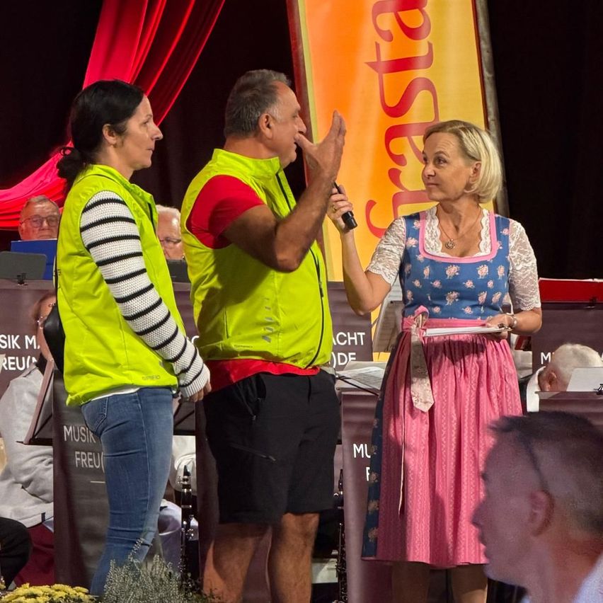 Drei Personen stehen auf einer Bühne, einer hält ein Mikrofon. Eine Frau in einem rosa Kleid hält ein Papier, und ein Mann in einem gelben Weste gestikuliert. Hinter ihnen steht ein Banner mit der Aufschrift 'Gasthaus'.