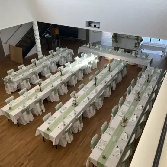 Ein Blick von oben auf einen großen Raum mit Tischen, die für eine besondere Veranstaltung vorbereitet sind. Die Tische sind mit weißen Tischdecken bedeckt, geschmückt mit grünen Bändern. Der Raum hat Holzböden und weiße Wände.