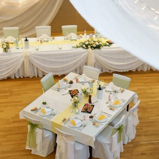 Ein gut dekorierter Bankettsaal mit zwei langen Tischen. Jeder Tisch hat Teller, Besteck, Weingläser und Blumen. Die Stühle sind mit weißer Stoff bedeckt. Der Boden ist aus Holz.