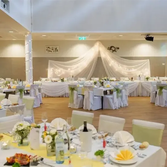 Ein Bankettsaal mit Tischen und Stühlen, alle in Weiß und Grün gekleidet. Jeder Tisch hat eine Weinflasche, Blumen und einen Teller. Der Raum hat Holzböden und weiße Wände. Über den Tischen befindet sich ein weißer Vorhang mit Lichtern.
