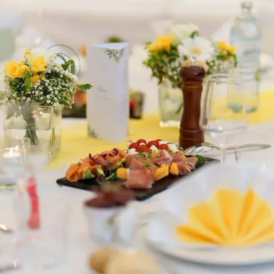 Eine Tischdekoration mit gelber Tischdecke, Blumen und einem Teller mit Tomaten, Käse und Fleisch. Es gibt Weingläser, Salzstreuer und eine Kerze.