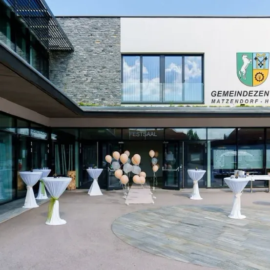 Ein modernes Gemeindezentrum mit einer Steinmauer außen. Ein Logo befindet sich über dem Eingang. Tische und Stühle sind draußen aufgestellt, mit Luftballons und einem Teppichboden.