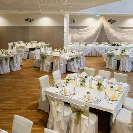 Ein Hochzeitsempfangsbereich mit weißen Tischdecken, weißen Stühlen und grünen Bändern. Eine weiße Decke mit Lichtern ist an der Decke. Jeder Tisch hat eine Blumenvase, Teller, Gläser und Besteck.