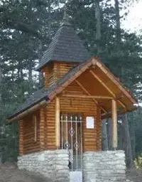 Eine kleine Holzkapelle mit schwarzem Dach und Steinfundament, umgeben von hohen Bäumen und Grün. Sie hat ein Metalltor und ein weißes Schild.