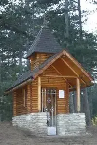 Eine kleine Holzkapelle mit schwarzem Dach und Steinfundament, umgeben von hohen Bäumen und Grün. Sie hat ein Metalltor und ein weißes Schild.