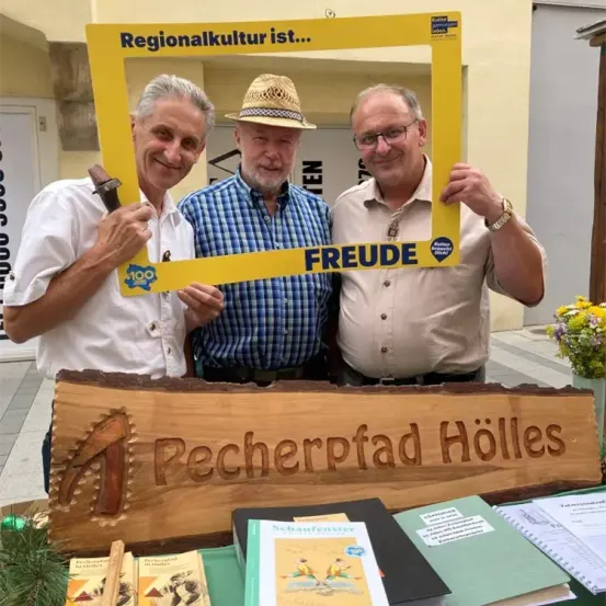 Drei Männer stehen hinter einem Holzschild mit der Aufschrift 'Pecherrpfad Holles' und halten einen gelben Rahmen mit der Aufschrift 'Regionalkultur ist... FREUDE'. Dahinter befindet sich ein Gebäude mit einem Schild und einer Blumenarrangement.