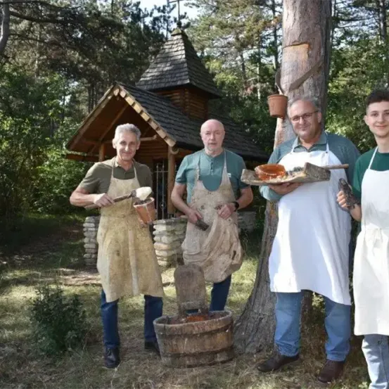 Vier Männer in Schürzen stehen in einem Wald in der Nähe einer Holzstruktur, einer hält ein Holzobjekt und ein anderer eine Holzschale mit Essen.