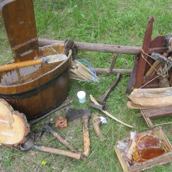 Ein hölzerner Eimer mit Griff steht auf Gras mit einigen verstreuten Holzwerkzeugen. Ein hölzerner Stuhl und eine Leiter sind ebenfalls in der Nähe. Eine hölzerne Kiste enthält einige Gegenstände.