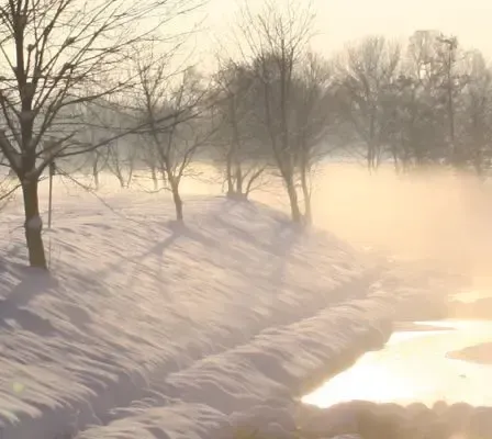 Eine verschneite Landschaft mit einem Fluss, der durch sie fließt, umgeben von laublosen Bäumen. Nebel steigt aus dem Fluss auf, und der Himmel ist klar und hell.