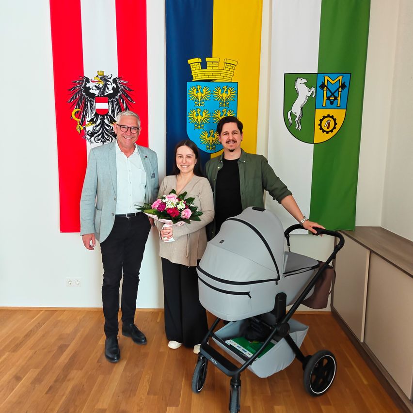 Drei Menschen stehen vor drei Flaggen. Ein Mann hält Blumen, eine Frau einen Kinderwagen und ein Mann im grünen Mantel hält den Griff des Kinderwagens. Sie alle lächeln.