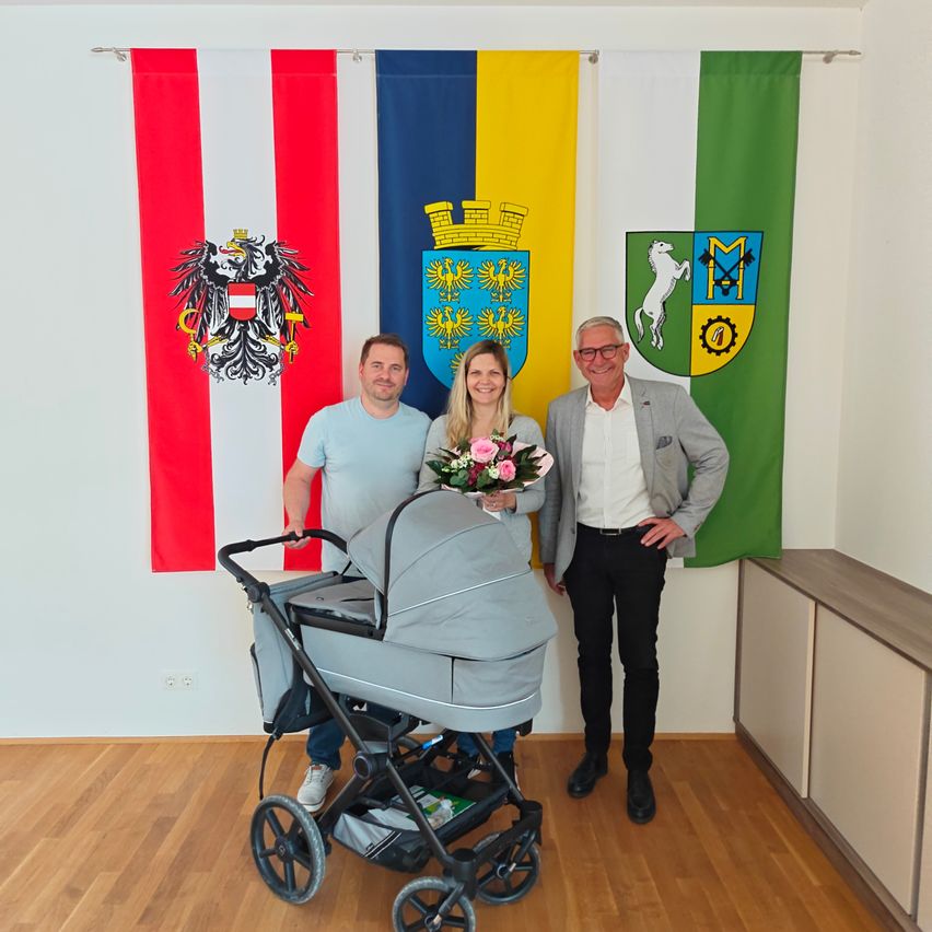 Drei Personen posieren vor einer mit Flaggen geschmückten Wand, darunter ein österreichisches Wappen. Zwei Männer und eine Frau stehen neben einem Kinderwagen.
