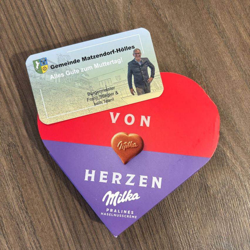 Eine herzförmige Schachtel Milka-Schokolade liegt auf einer Holzoberfläche, mit einer Karte mit einem Mann und Text darüber.