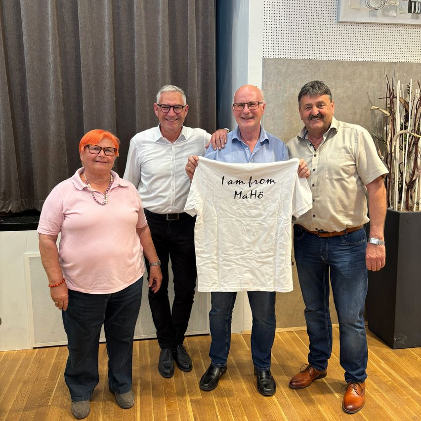 Vier Personen stehen lächelnd zusammen und halten ein weißes T-Shirt mit der Aufschrift 'Ich komme aus Mahó'. Die beiden Männer tragen jeweils eine Brille, einer von ihnen auch eine Uhr. Die Frau trägt eine Halskette und hat orangefarbenes Haar. Hinter ihnen befindet sich ein Vorhang und eine Pflanze in einem Topf.