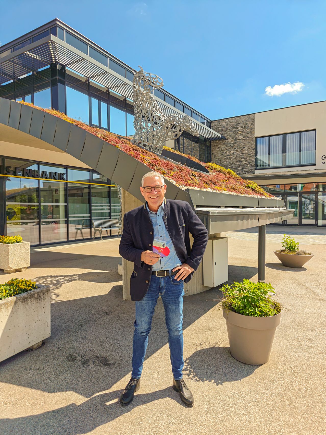 Ein Mann steht vor einem Gebäude mit grünem Dach, trägt eine Brille und hält eine Broschüre in der Hand. In der Nähe stehen Topfpflanzen und eine Bank.