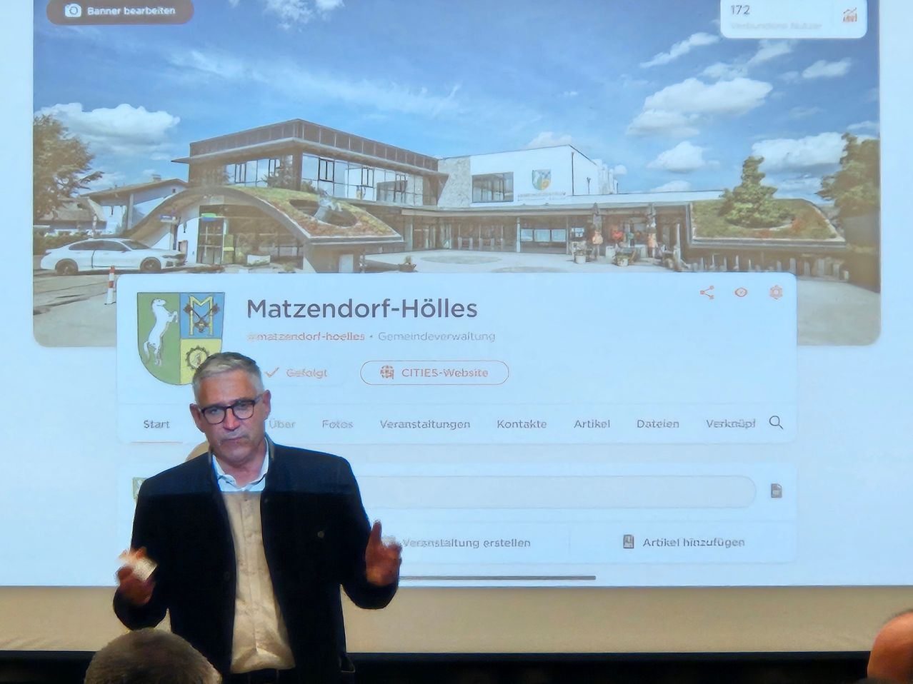 Ein Mann hält vor einem Bildschirm eine Präsentation über die Matzendorf-Hölles-Website. Das Gebäude hat ein grünes Dach, und die Präsentation enthält Optionen wie Artikel, Fotos, Veranstaltungen und Kontaktinformationen.
