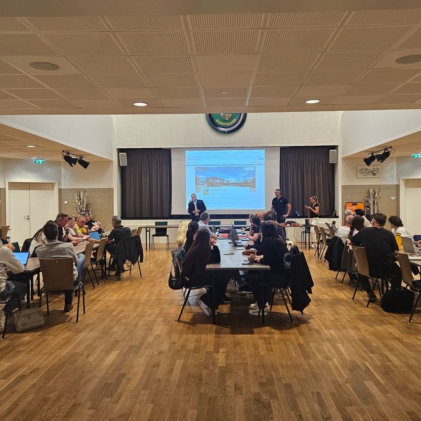 Ein Seminar mit Teilnehmern in einer Halle mit Holzboden. Zwei Männer stehen an einem Tisch und halten eine Präsentation mit einem Projektor ab.