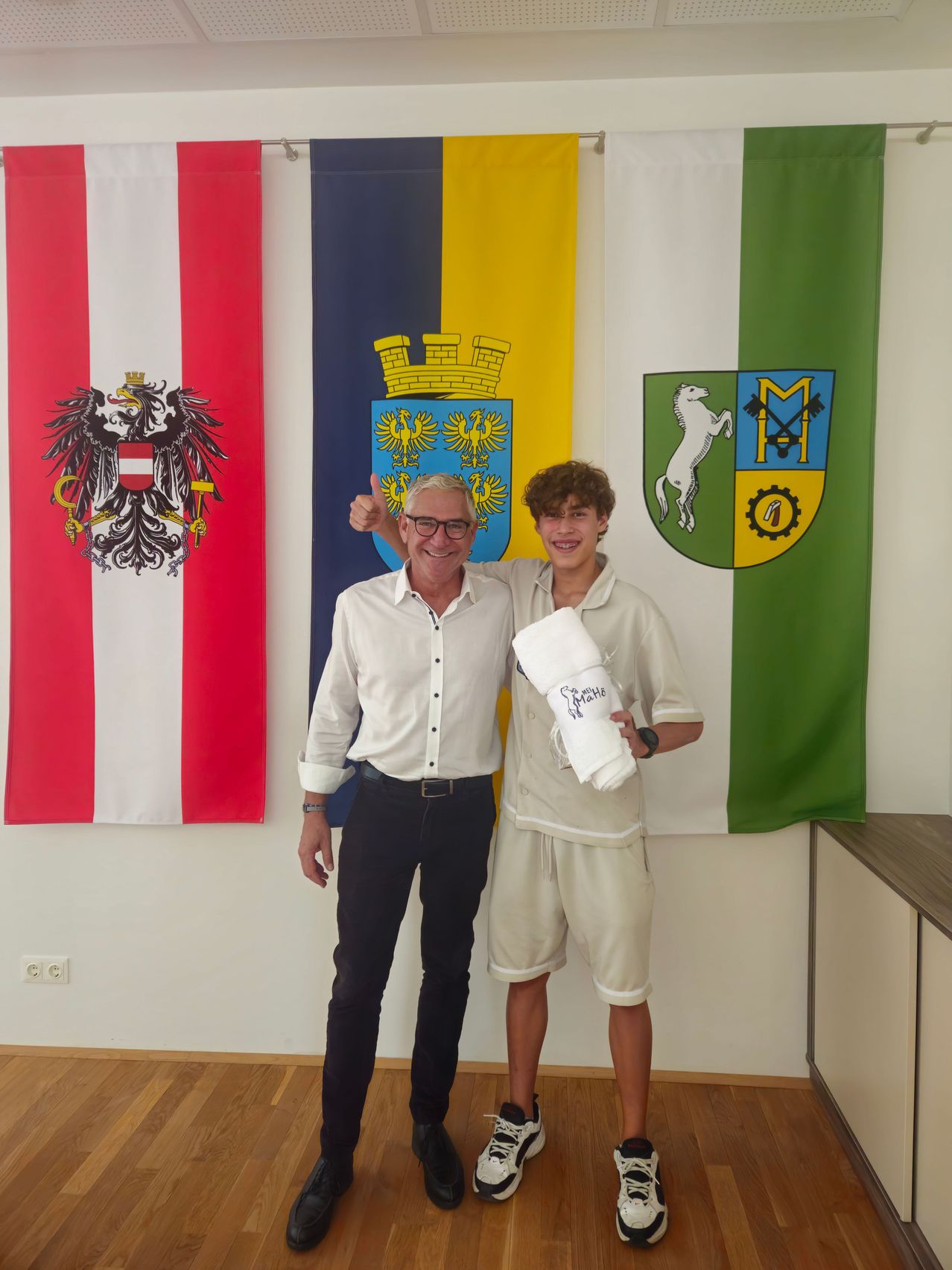Zwei Männer stehen vor einer Wand mit drei Bannern. Der Mann auf der linken Seite gibt einen Daumen hoch, während der Mann auf der rechten Seite ein aufgerolltes weißes Tuch hält.