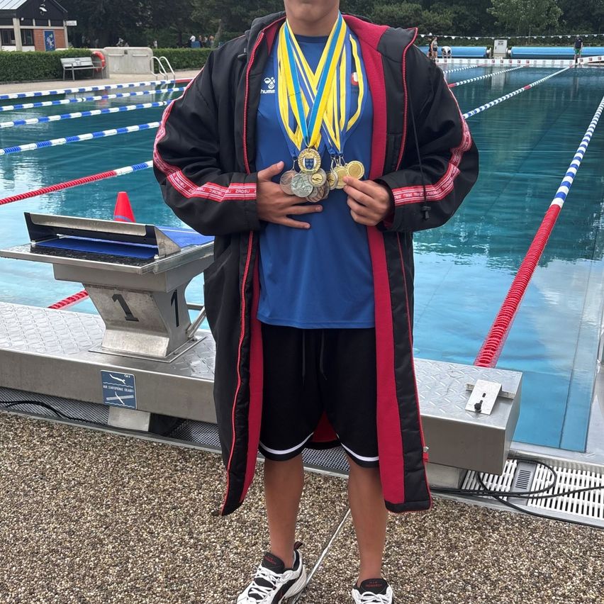 Ein junger Mann steht am Rand eines Schwimmbeckens, trägt ein blaues T-Shirt und mehrere goldene Medaillen um den Hals. Er ist in einer schwarz-roten Jacke mit weißen und roten Besätzen gekleidet.