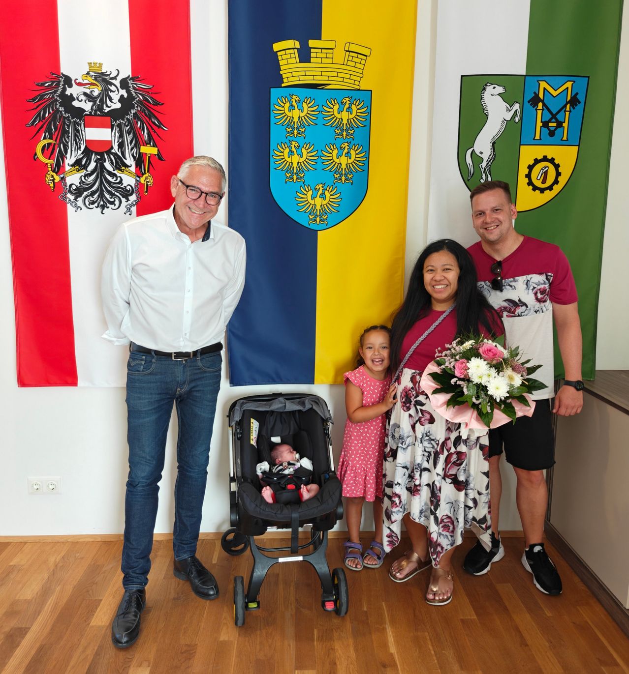 Eine Familie steht vor einer Wand mit drei verschiedenen Flaggen. Der Mann mit Brille trägt ein weißes Hemd, während die Frau Blumen hält. Sie posieren mit einem Baby im Kinderwagen und einem kleinen Mädchen.