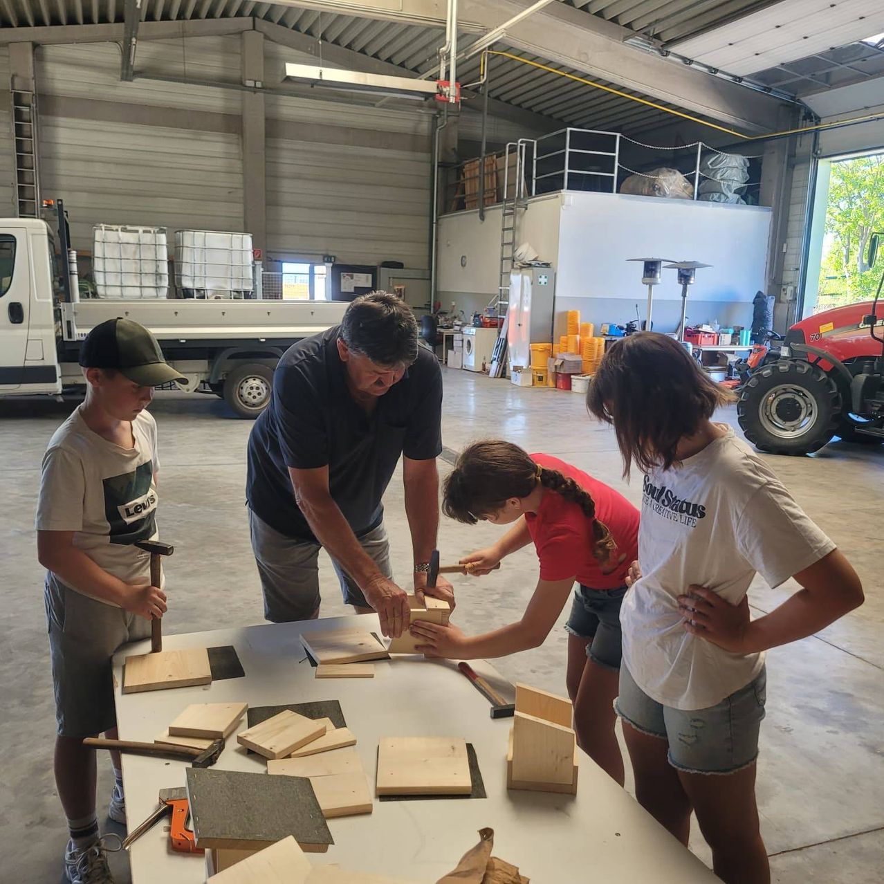 Ein Mann und drei Kinder arbeiten in einer Garage an einem Holzprojekt. Sie haben Holzteile, Werkzeuge und einen Tisch aufgebaut. Im Hintergrund stehen ein LKW, eine Leiter und verschiedene Gegenstände.