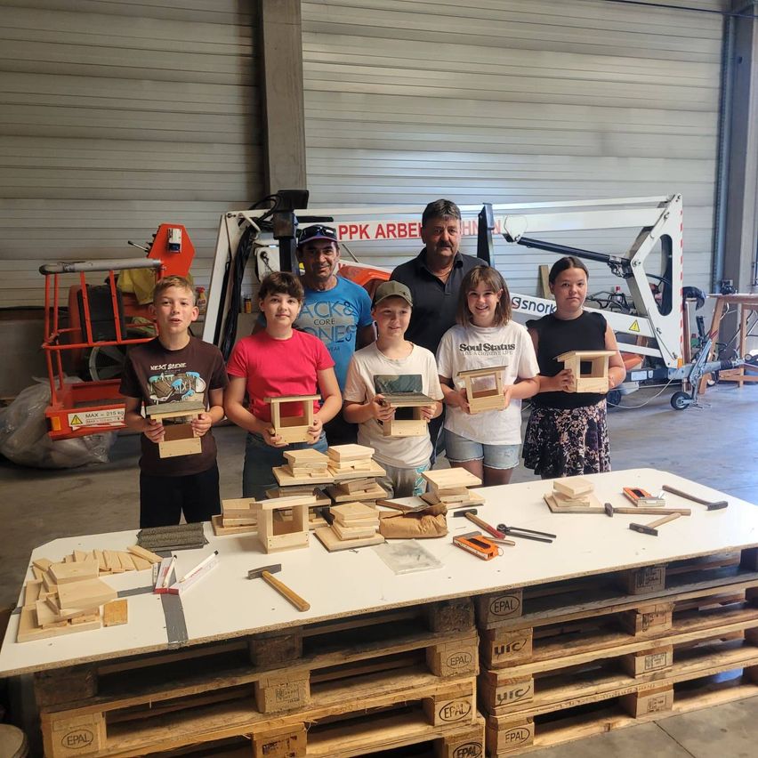 Eine Gruppe von Kindern und Erwachsenen in einer Werkstatt, die Holzvogelhäuschen halten. Ein Tisch voller Holzstücke und Werkzeuge. Ein Gabelstapler im Hintergrund.